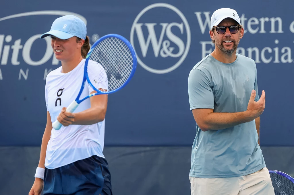 Iga Świątek i Wim Fissette podczas turnieju WTA 1000 w Cincinnati Dwie osoby na korcie tenisowym, jedna z nich trzyma rakietę i jest ubrana w sportowy strój oraz czapkę, druga osoba w jasnej koszulce i okularach przeciwsłonecznych uśmiecha się i wykonuje gest ręką, w tle niebieska tablica z logotypami.