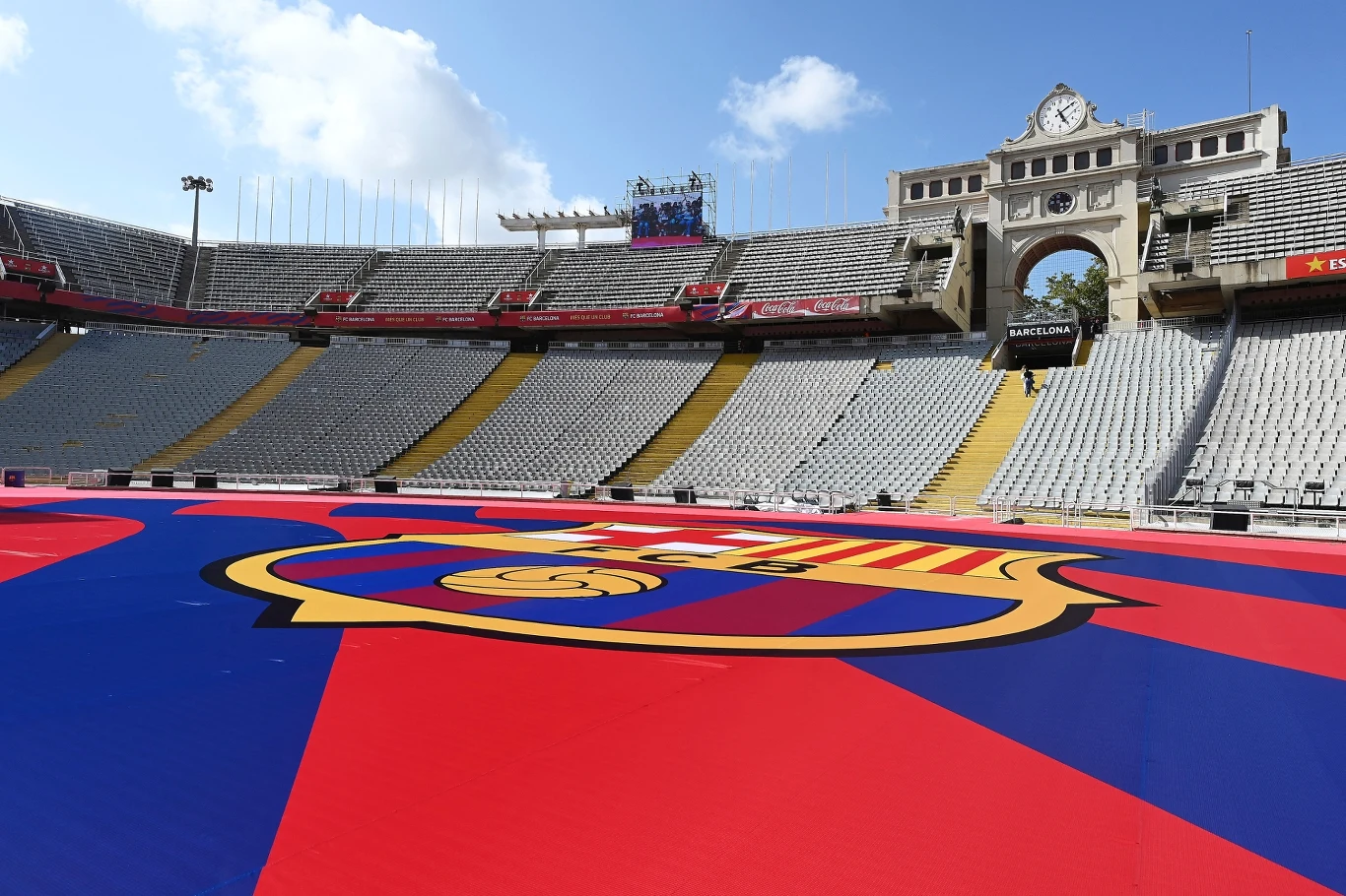 Rozległa panorama pustego stadionu z widocznym ogromnym herbem FC Barcelona rozłożonym centralnie na murawie, w tle historyczna trybuna z zegarem oraz błękitne niebo z chmurami.
