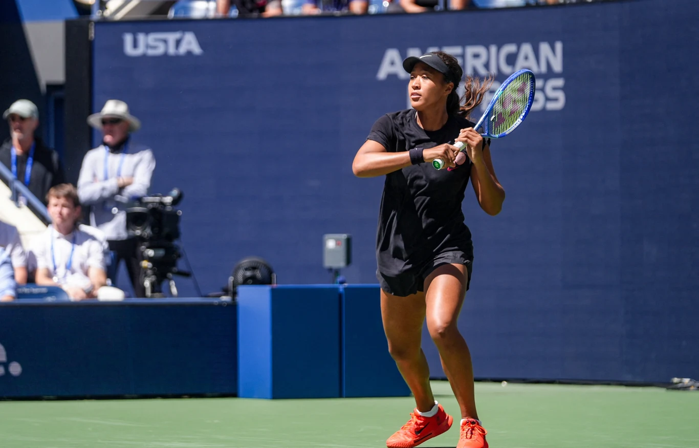 Naomi Osaka podczas miksta na US Open Tenisistka w czarnym stroju przygotowuje się do uderzenia piłki na profesjonalnym korcie, w tle widoczni są sędziowie oraz tablica turnieju US Open.