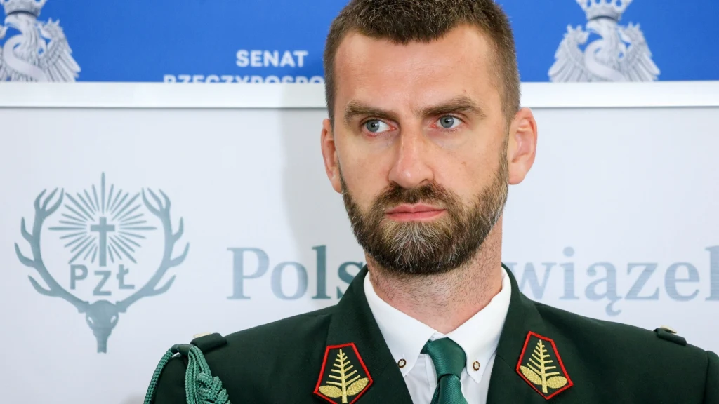Marcin Możdżonek Mężczyzna w zielonym mundurze z odznakami łowieckimi stoi na tle planszy z logotypem Polskiego Związku Łowieckiego oraz napisem 'Senat Rzeczypospolitej Polskiej'.