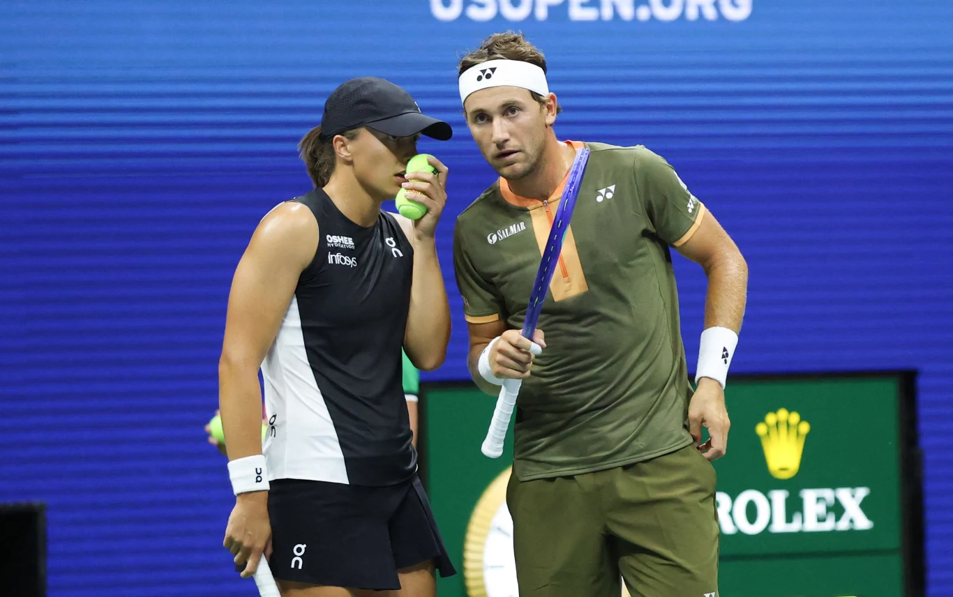 Dwoje tenisistów w trakcie rozmowy na korcie, kobieta w czarno-białym stroju trzyma piłkę tenisową przy twarzy, mężczyzna w oliwkowym stroju z rakietą opiera się na biodrze, w tle widoczne logo turnieju oraz zegar Rolex.