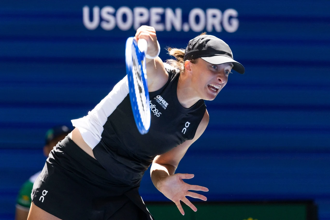 Iga Świątek Tenisistka w czarnej koszulce i czapce w trakcie dynamicznego serwisu podczas turnieju US Open, z widocznym niebieskim kortem i logo USOPEN.ORG w tle.
