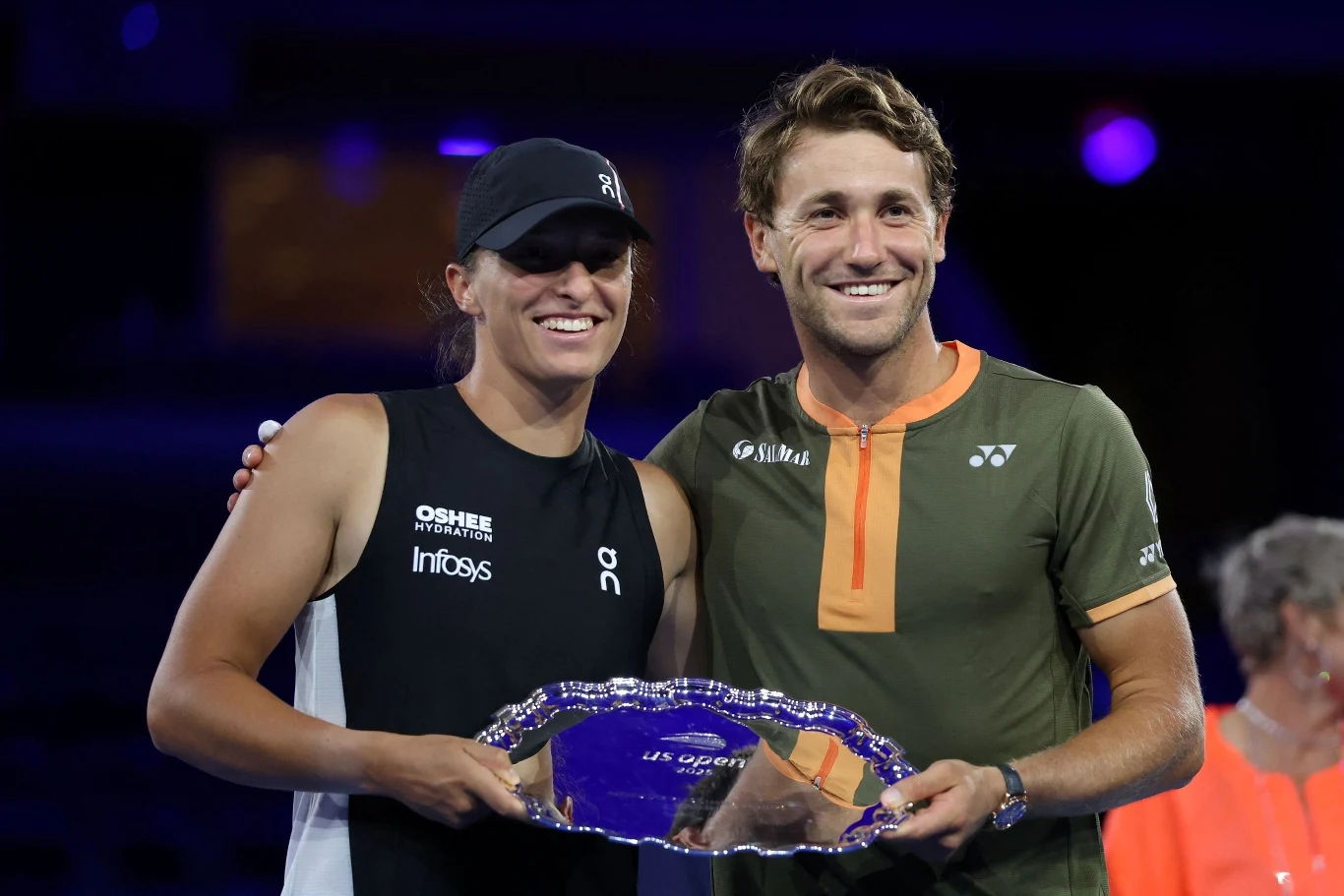 Dwoje sportowców stoi razem i uśmiecha się do kamery, jedna z osób trzyma w rękach srebrną tacę będącą nagrodą za osiągnięcie sportowe, obie osoby ubrane są w stroje sportowe.