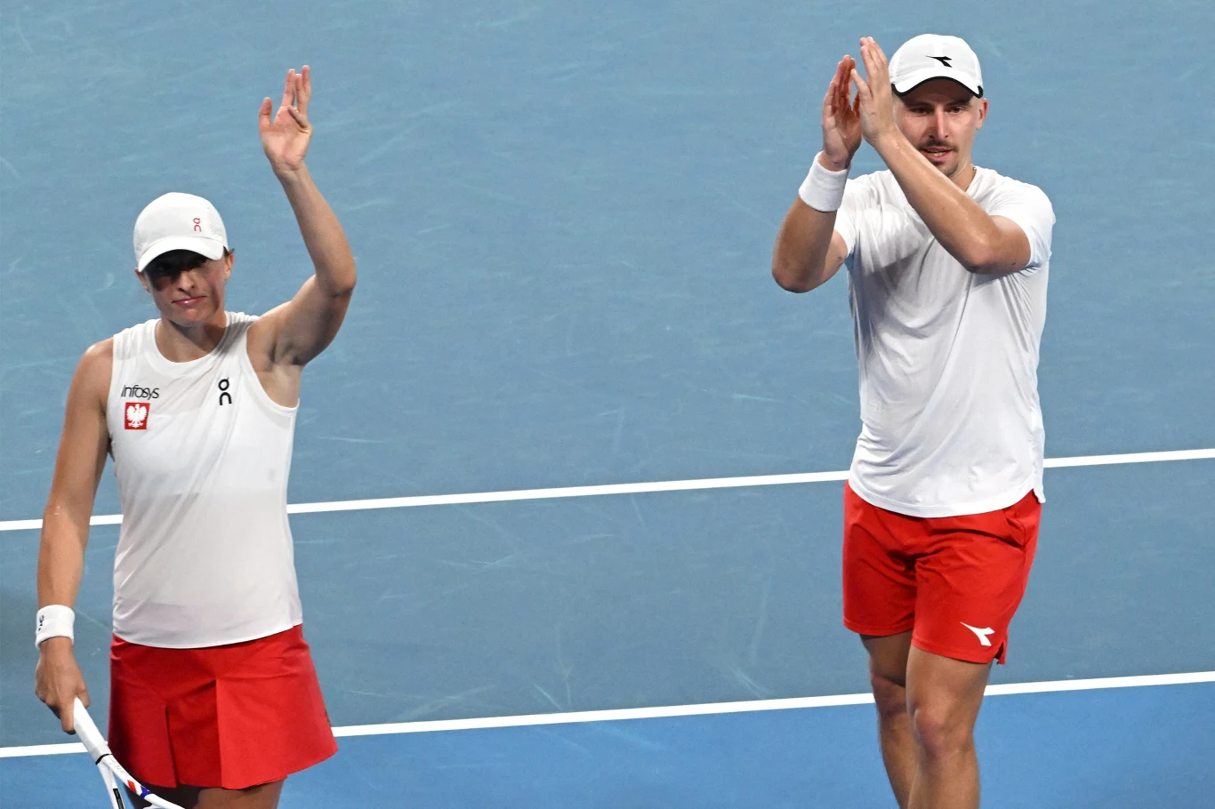 Dwójka tenisistów w białych koszulkach i czerwonych spodenkach stoi na korcie tenisowym, oboje unoszą jedną rękę w geście powitania lub podziękowania, w tle niebieska nawierzchnia kortu.