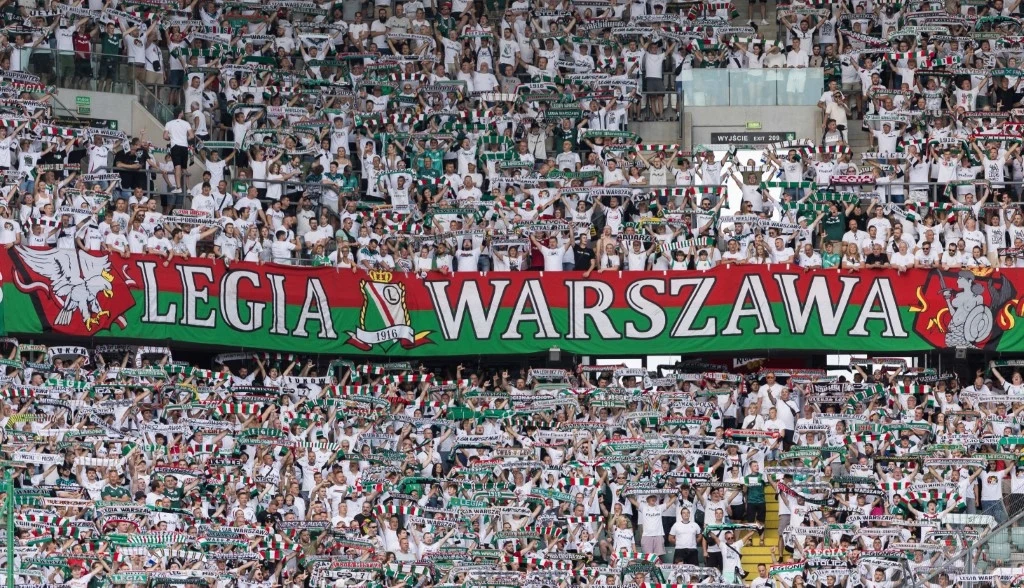 Duży tłum kibiców ubranych w białe koszulki na stadionie, trzymających szaliki i transparent z napisem Legia Warszawa, w tle dominuje kolorystyka biało-zielono-czerwona.