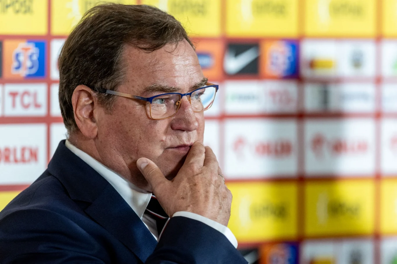 Jan Urban Mężczyzna w garniturze i okularach stoi na tle ścianki sponsorskiej, wygląda na zamyślonego, opierając rękę o twarz.