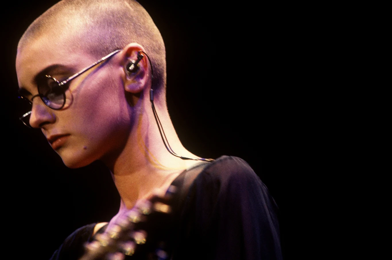 Sinead O'CONNOR Młoda osoba z ogoloną głową i okularami przeciwsłonecznymi, nosząca czarną bluzkę, z mikrofonem dousznym i skupionym wyrazem twarzy, sfotografowana na tle ciemnego tła.