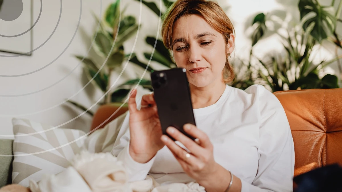 Emitel Kobieta o jasnych włosach w białej bluzce siedzi na kanapie i trzyma w dłoni smartfon, patrząc na ekran, w tle rośliny doniczkowe