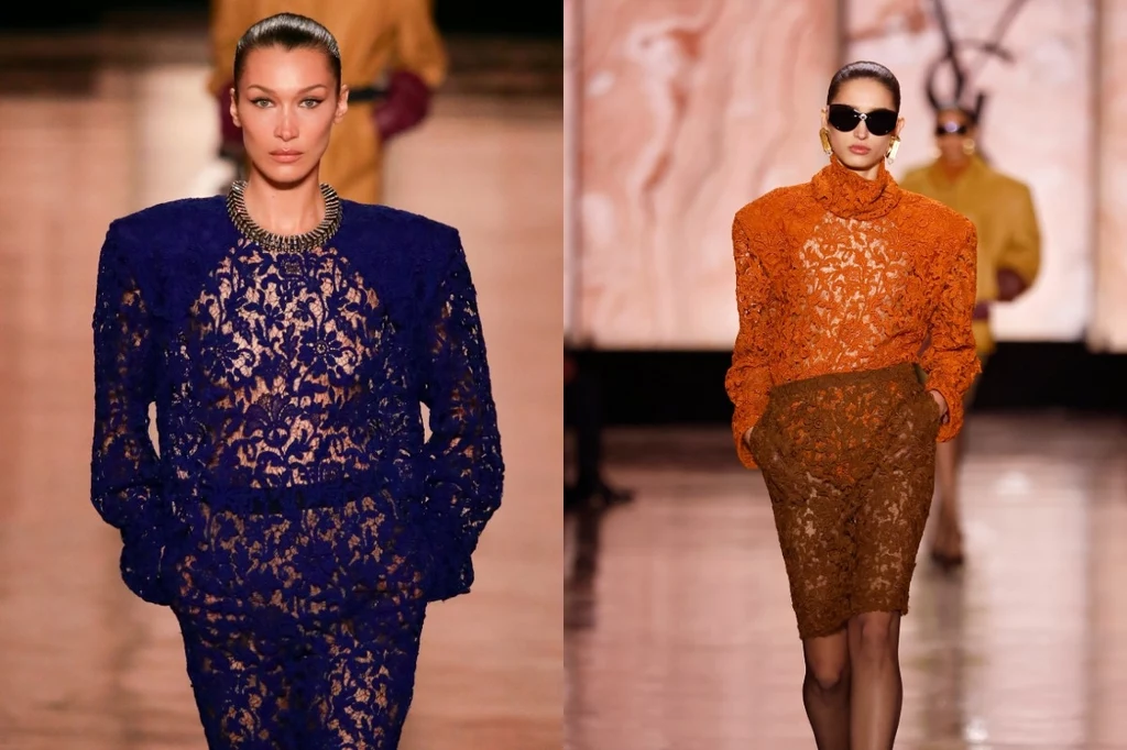 Saint Laurent 2025-2026 Dwie modelki prezentujące eleganckie, koronkowe stylizacje na wybiegu, jedna ubrana w granatowy komplet z szerokimi ramionami, druga w pomarańczowy zestaw z przezroczystej tkaniny i spódnicą za kolano, w tle rozmazany widok reszty pokazu mody.