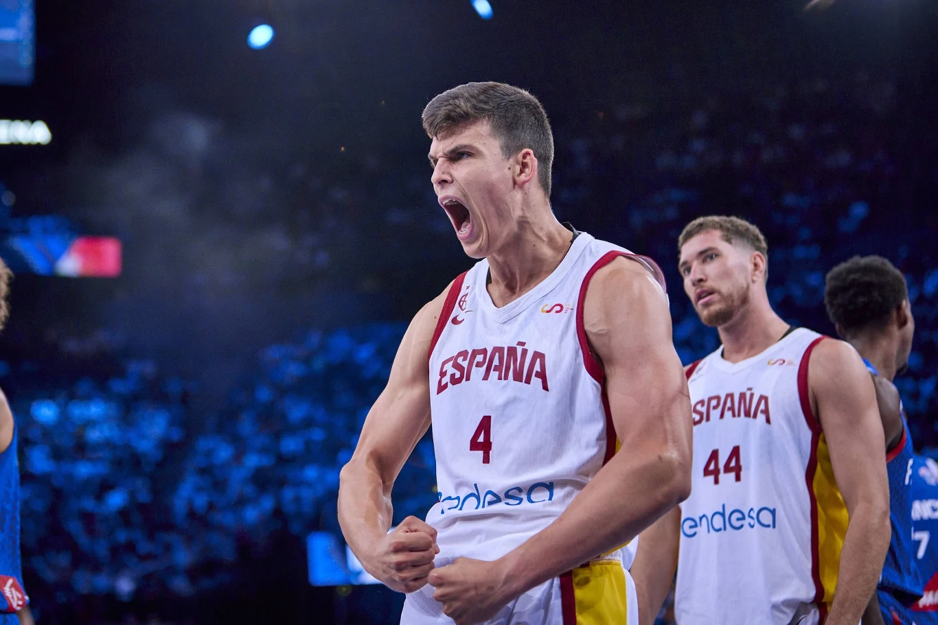 Koszykarz w stroju reprezentacji Hiszpanii wyraża silne emocje, stojąc z zaciśniętymi pięściami na tle hali sportowej, obok niego widać innego zawodnika tej samej drużyny.