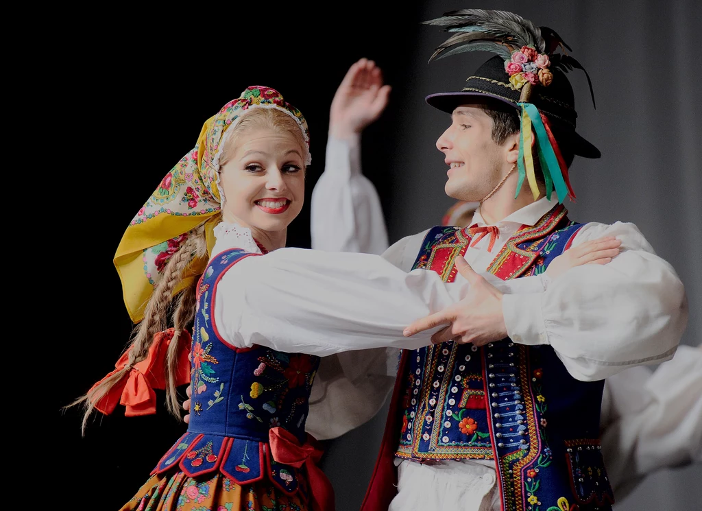Para młodych ludzi ubrana w tradycyjne polskie stroje ludowe tańczy, kobieta wzbogaca swój strój o kolorową chustę na głowie i bogato haftowany gorset, mężczyzna nosi kapelusz z piórem oraz ozdobną kamizelkę.