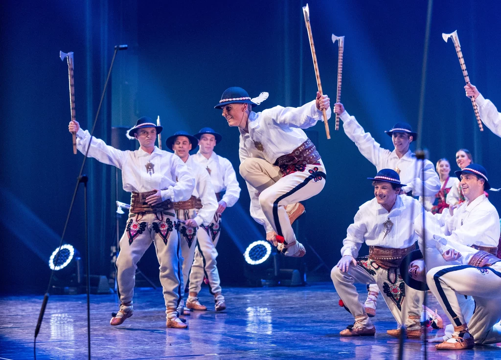 Grupa tancerzy w tradycyjnych strojach góralskich wykonuje skoczny taniec na scenie, część z nich trzyma ozdobne ciupagi, a jeden z tancerzy jest w trakcie skoku.