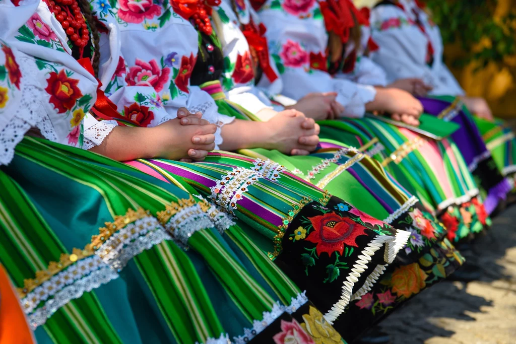 Kolorowe, tradycyjne stroje ludowe z kwiatowymi haftami, koralikami i bogato zdobionymi spódnicami, ubrane przez siedzące kobiety trzymające dłonie na kolanach.