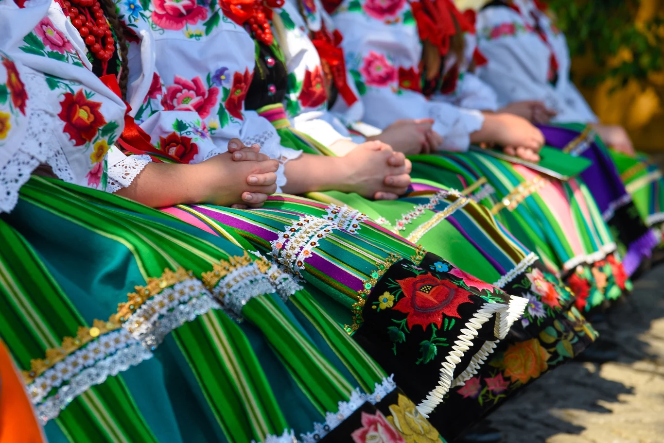 Regionalne, ludowe stroje są pełne barw i kolorów Kolorowe, tradycyjne stroje ludowe z kwiatowymi haftami, koralikami i bogato zdobionymi spódnicami, ubrane przez siedzące kobiety trzymające dłonie na kolanach.