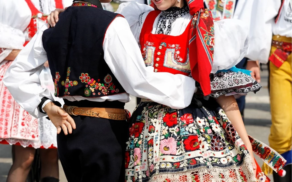 Para ubrana w tradycyjne stroje ludowe tańczy, zwracając uwagę na bogato zdobione hafty, kolorowe aplikacje na spódnicy i kamizelce, wyraźnie widoczne detale folklorystycznego ubioru podkreślającego regionalny charakter wydarzenia.