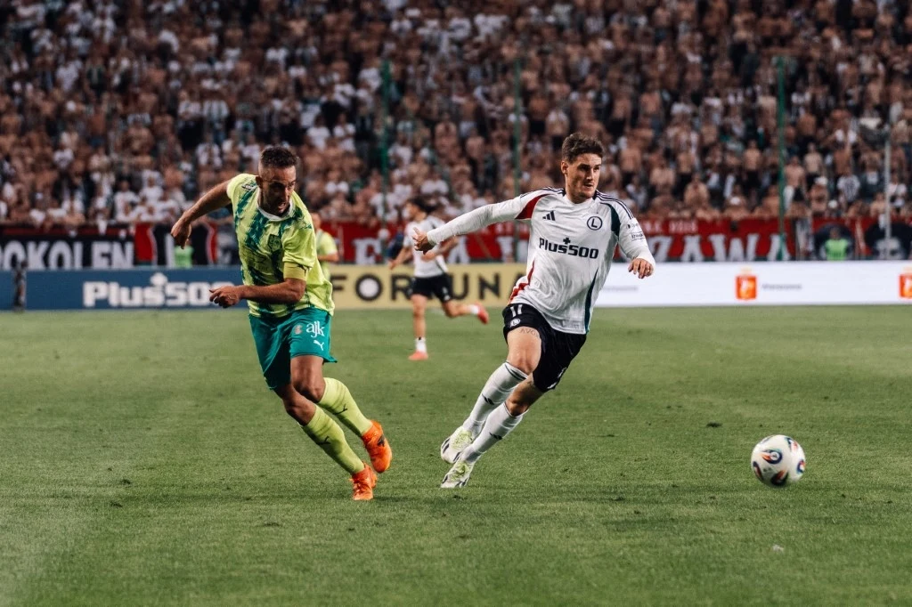 Dwóch piłkarzy rywalizuje o piłkę na stadionie przy pełnych trybunach, jeden w białym stroju, drugi w zielonym, w tle widać kibiców i reklamy na bandzie.