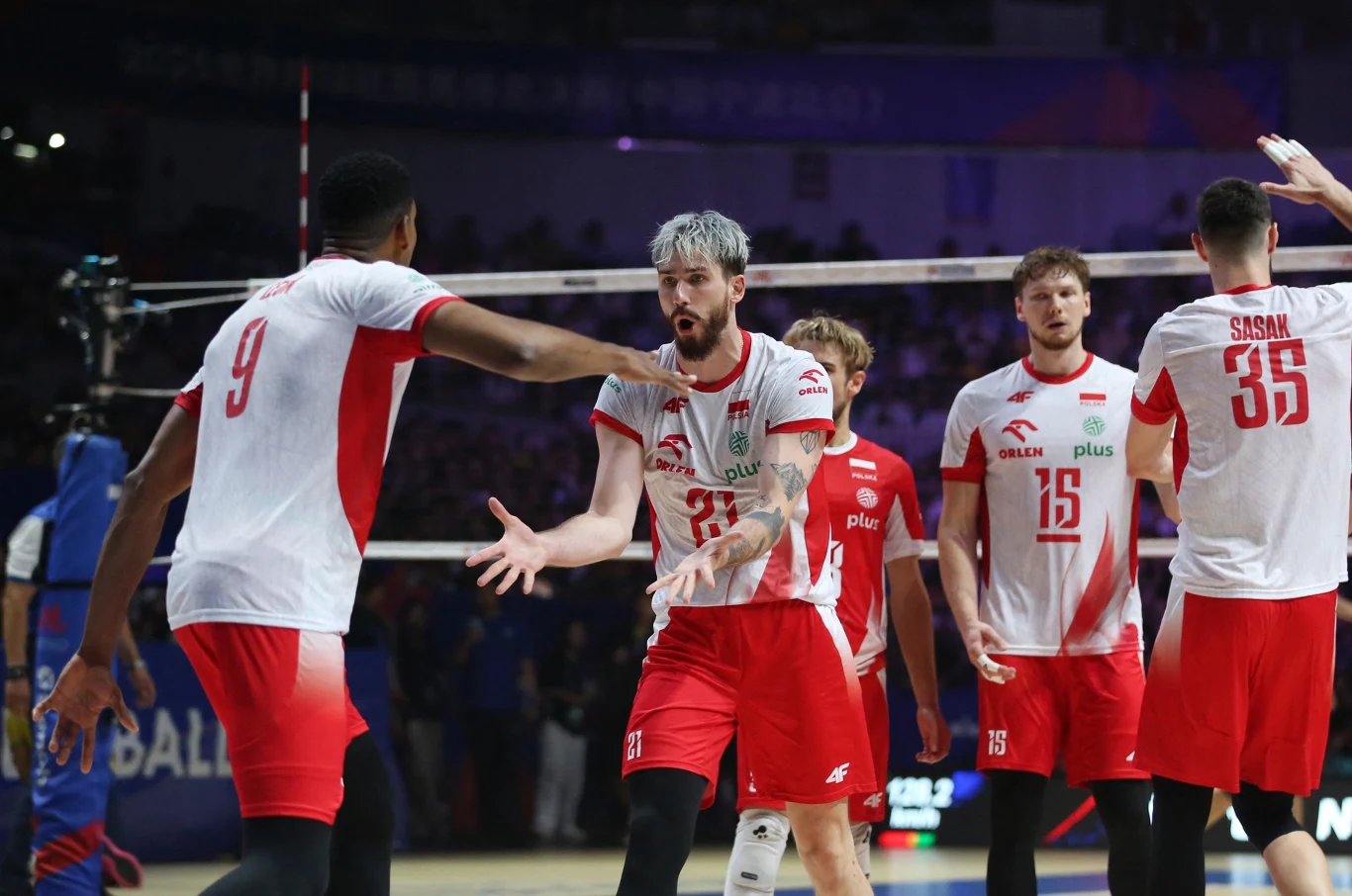 Siatkarze polskiej reprezentacji w dynamicznym momencie meczu, zawodnicy ubrani w białe i czerwone stroje, wykonują gesty współpracy i zaangażowania, w tle widownia na trybunach oraz siatka.