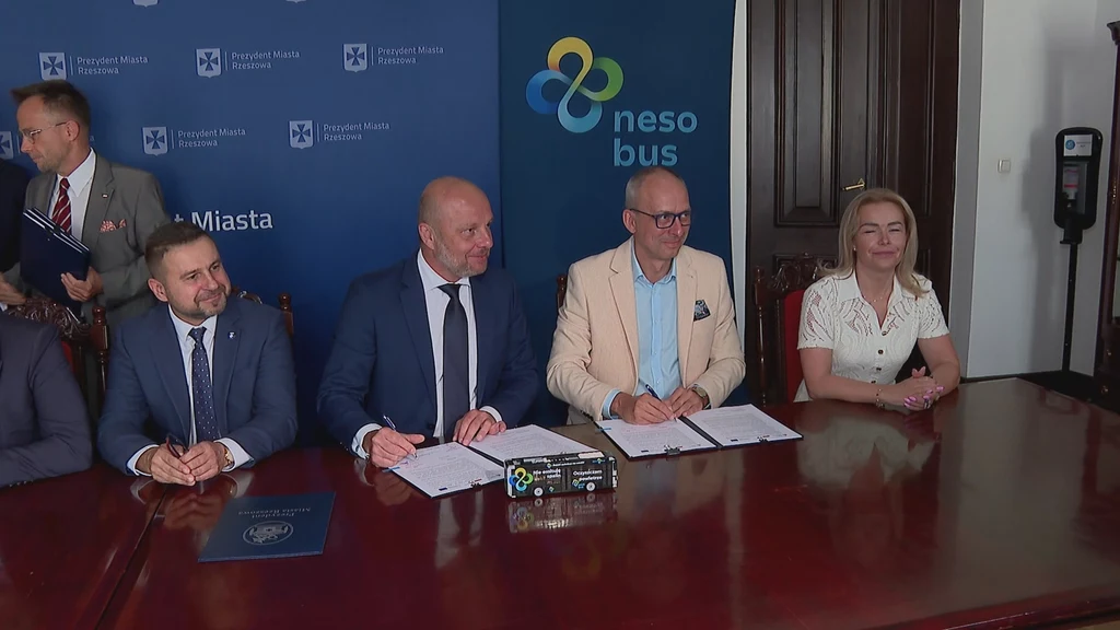 W Rzeszowie podpisano umowę na dostawę 20 autobusów wodorowych NesoBus Cztery osoby siedzą przy dużym, drewnianym stole i podpisują dokumenty, w tle niebieskie tło z logotypami oraz banerem firmy nesobus, atmosfera oficjalna, eleganckie ubiory uczestników.