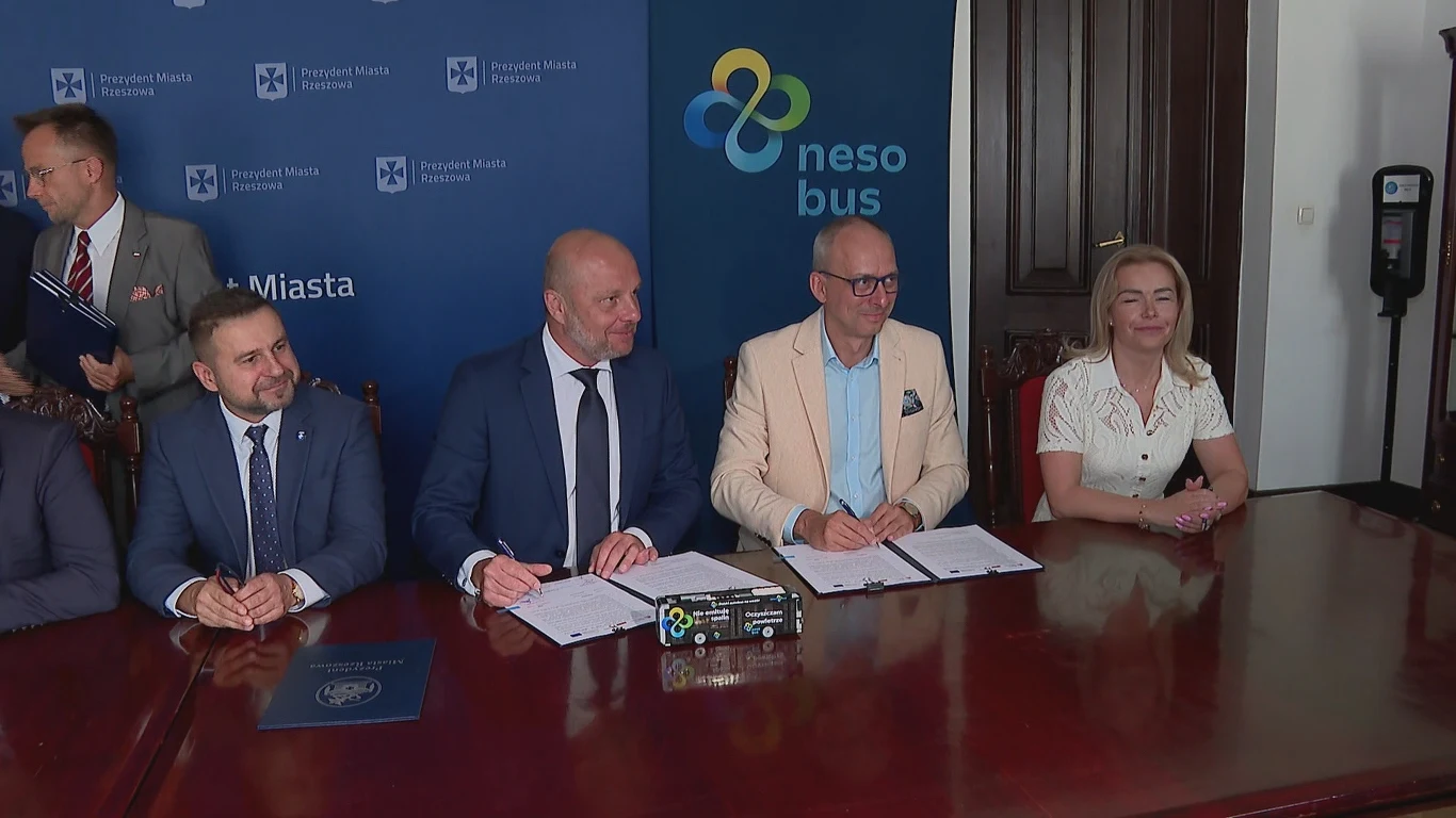 W Rzeszowie podpisano umowę na dostawę 20 autobusów wodorowych NesoBus Cztery osoby siedzą przy dużym, drewnianym stole i podpisują dokumenty, w tle niebieskie tło z logotypami oraz banerem firmy nesobus, atmosfera oficjalna, eleganckie ubiory uczestników.
