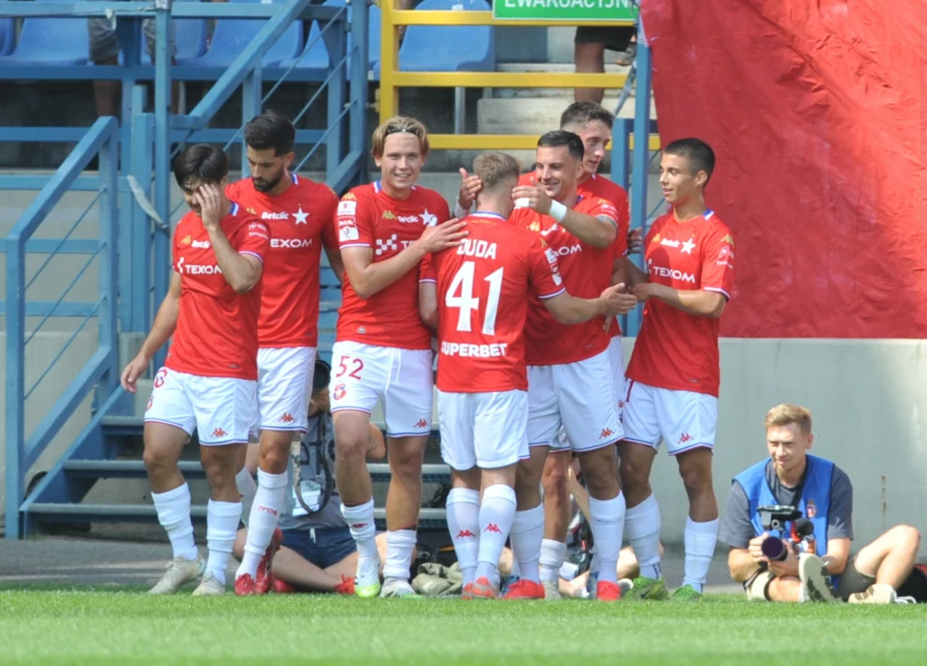 Wisła Kraków Grupa piłkarzy w czerwonych koszulkach cieszy się z bramki na murawie stadionu, wokół nich widoczni kibice na trybunach oraz fotograf na skraju boiska.