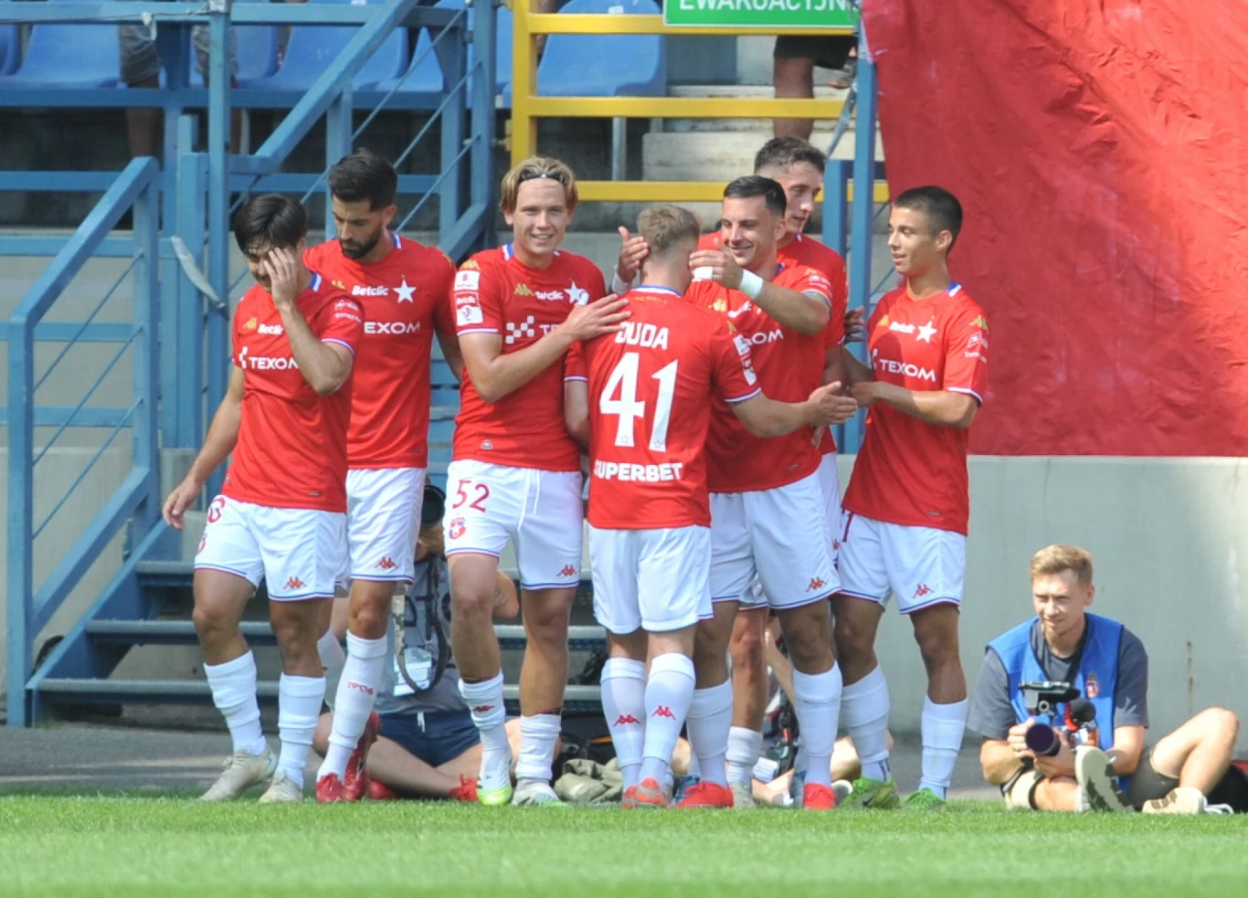 Grupa piłkarzy w czerwonych koszulkach cieszy się z bramki na murawie stadionu, wokół nich widoczni kibice na trybunach oraz fotograf na skraju boiska.