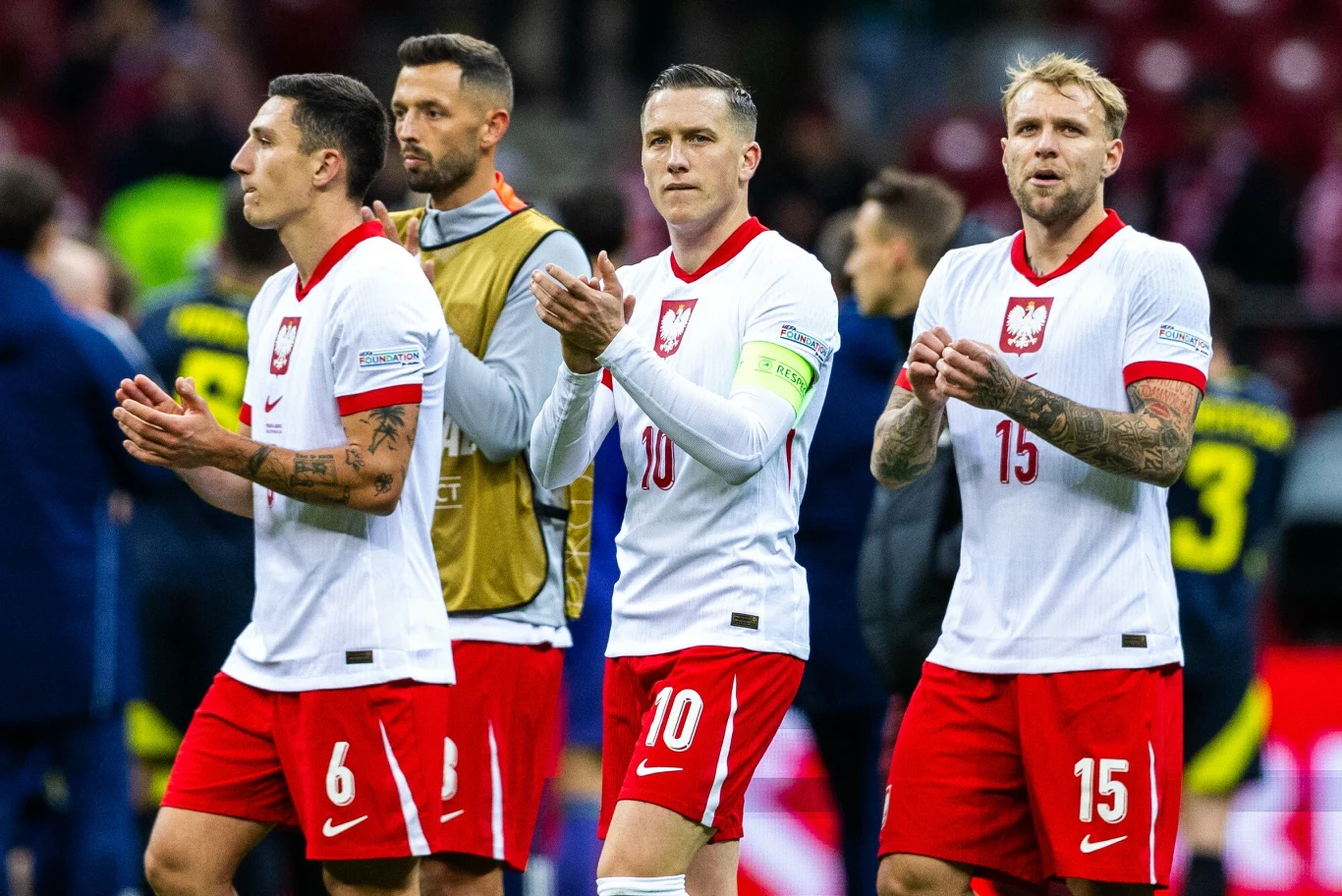 Polscy piłkarze w oficjalnych strojach reprezentacji stoją blisko siebie i biją brawo, w tle rozmyci kibice i inni zawodnicy.