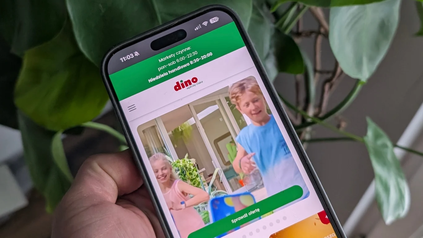 Dino jak Biedronka i Lidl. Będzie miało własną aplikację. Ekran smartfona prezentuje stronę internetową sieci handlowej Dino z widocznym zdjęciem uśmiechniętych dzieci w jasnych ubraniach na tle roślin i domowego wnętrza, całość trzymana jest w ręce na tle dużych zielonych liści.