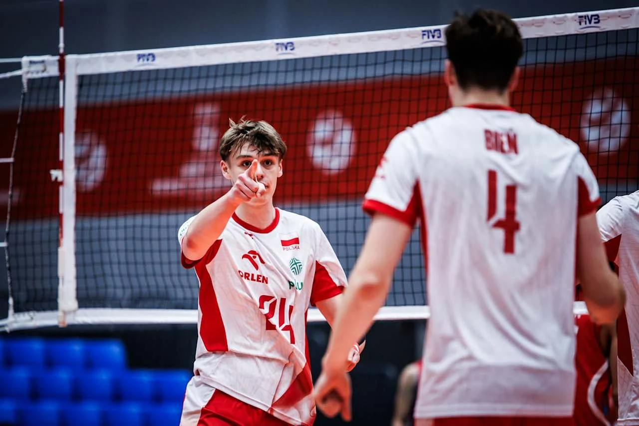 Dwóch siatkarzy w biało-czerwonych strojach drużyny narodowej znajduje się na boisku przy siatce, jeden z nich wskazuje palcem w kierunku kamery, drugi stoi bokiem z widocznym numerem 4 na plecach. W tle widoczna część hali sportowej.