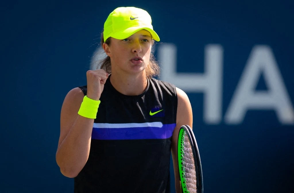 Iga Świątek zadebiutowała w US Open w 2019 roku Tenisistka w czapce i opasce w kolorze neonowej żółci, ubrana w czarną koszulkę z niebieskim paskiem, zaciśnięta pięść i wyraz determinacji na twarzy, w dłoni trzyma rakietę tenisową, w tle rozmazane litery na ciemnoniebieskim tle.