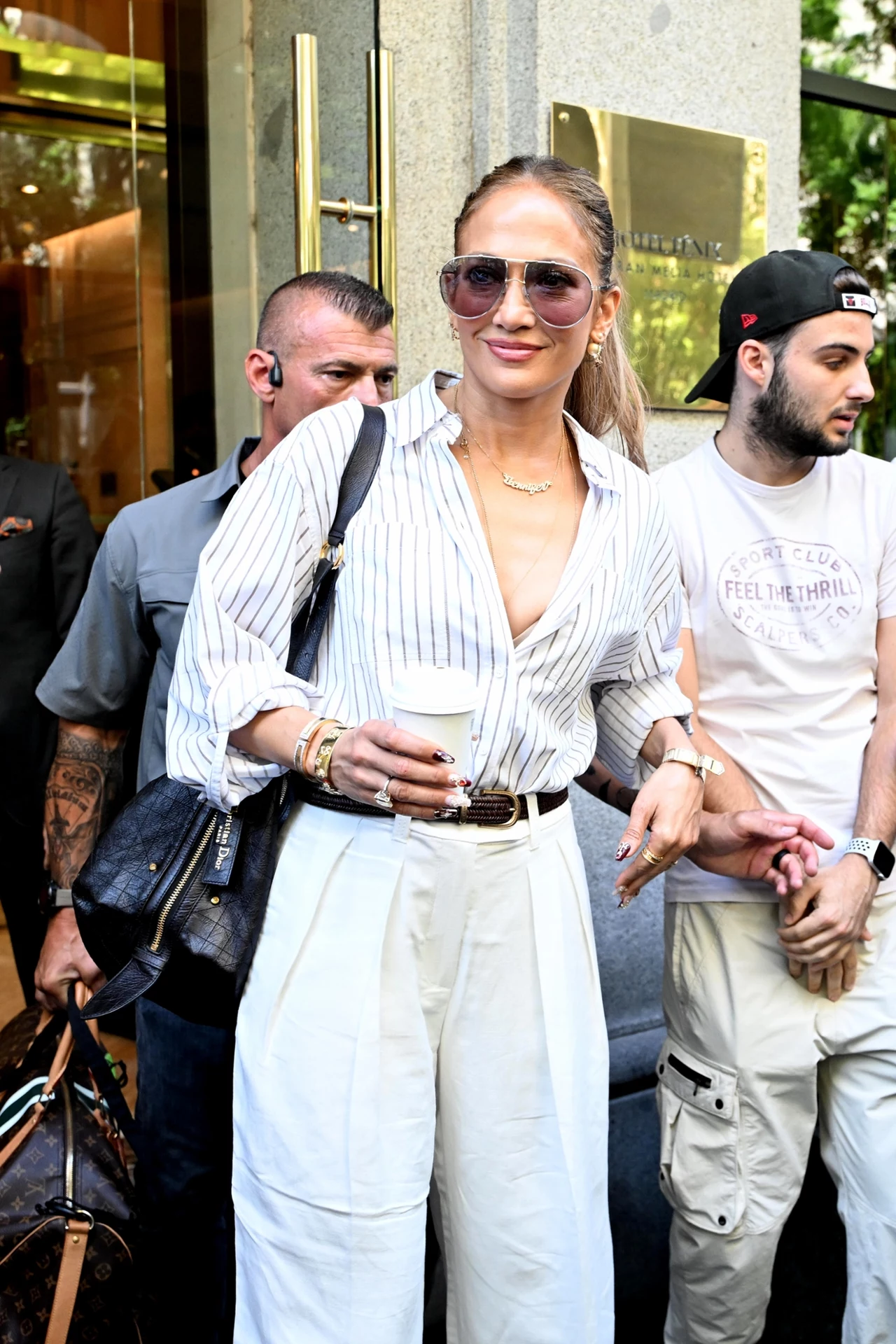 "Cichy luksus" w wydaniu Jennifer Lopez to strzał w dziesiątkę właśnie dzięki tym spodniom Kobieta w stylowym, letnim stroju stoi na ulicy w towarzystwie ochroniarzy i innych osób, trzymając w ręku papierowy kubek z napojem, ma na sobie białe szerokie spodnie, koszulę w paski, okulary przeciwsłoneczne i liczne bransoletki.