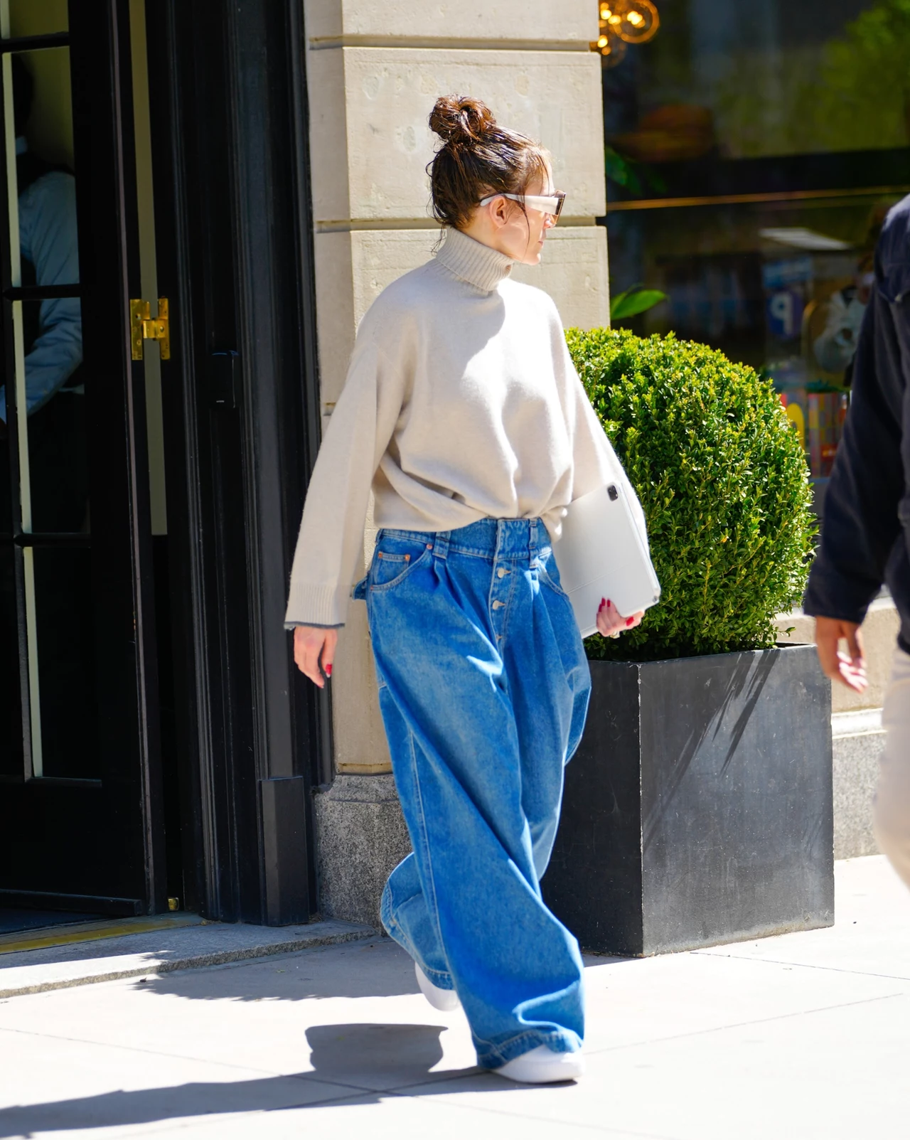 Szerokie jeansowe spodnie to podstawa luźnej garderoby Jennifer Lopez Kobieta ubrana w jasny, oversize'owy sweter z golfem oraz szerokie, niebieskie jeansy idzie chodnikiem w słoneczny dzień, z laptopem pod pachą, na tle zielonego krzewu i budynku.