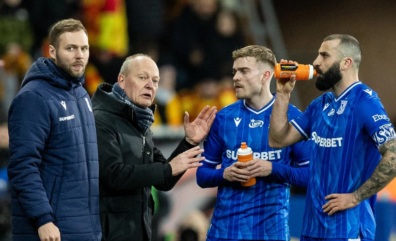 Trener rozmawia z piłkarzami drużyny podczas przerwy w meczu, jeden z zawodników pije z bidonu. Wszyscy ubrani są w niebieskie stroje sportowe, w tle widoczni są kibice na trybunach.