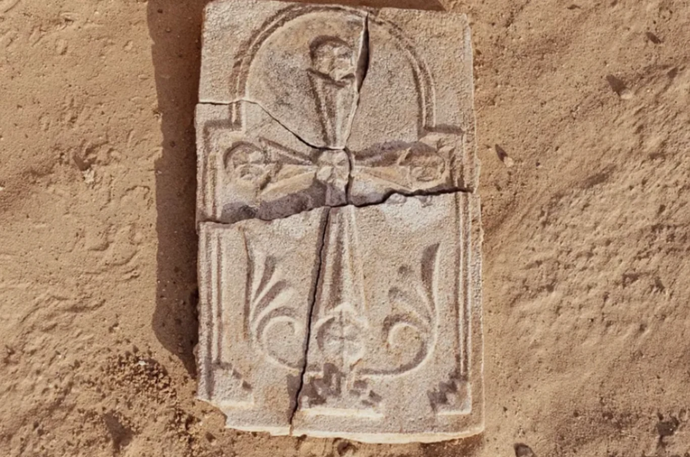 1400-letni krzyż odkryty w ZEA. Rzadkie świadectwo wiary chrześcijańskiej na Półwyspie Arabskim Kamienna płyta z wyrytym krzyżem i dekoracyjnymi motywami roślinnymi, uszkodzona w kilku miejscach, leżąca na piasku.