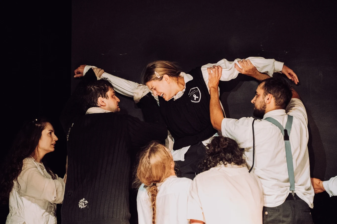 Grupa ludzi ubranych w teatralne kostiumy współpracuje, unosząc kobietę w ciemnym bezrękawniku na tle czarnej ściany, gestami i mimiką przedstawiają intensywne emocje.