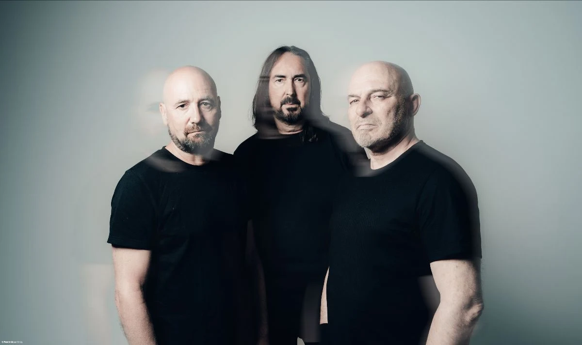 Coroner ujawnił szczegóły nowej płyty Trzech mężczyzn o poważnych minach stoi obok siebie w neutralnym, jasnym tle, ubrani w czarne koszulki, z lekkim efektem rozmycia na niektórych postaciach.