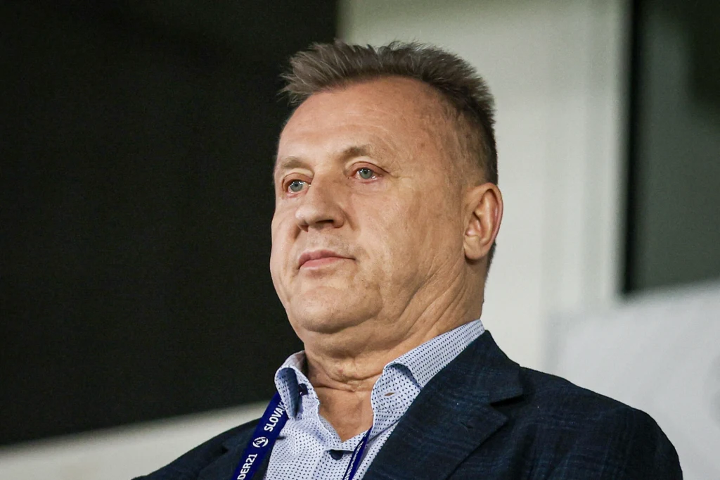 Mężczyzna w średnim wieku w eleganckim garniturze, z wyrazem skupienia i lekko zaciśniętymi ustami, stoi na tle nowoczesnego wnętrza, widoczny identyfikator zawieszony na szyi.