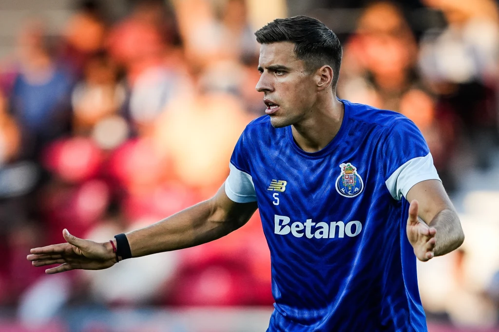 Piłkarz w niebieskiej koszulce z herbem klubu FC Porto wykonuje gest rękoma podczas meczu, tło rozmyte z widocznymi kibicami.