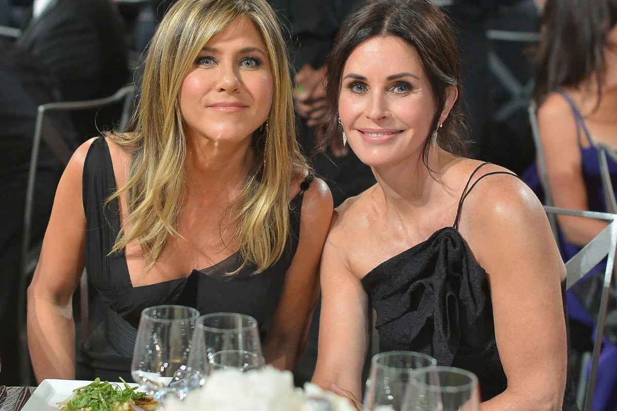 Jennifer Aniston i Courteney Cox spotkały się na podwójnej randce ze swoimi partnerami Dwie elegancko ubrane kobiety siedzące obok siebie przy stole na uroczystym wydarzeniu, przed nimi znajdują się kieliszki i talerze z jedzeniem, obie uśmiechają się do obiektywu.