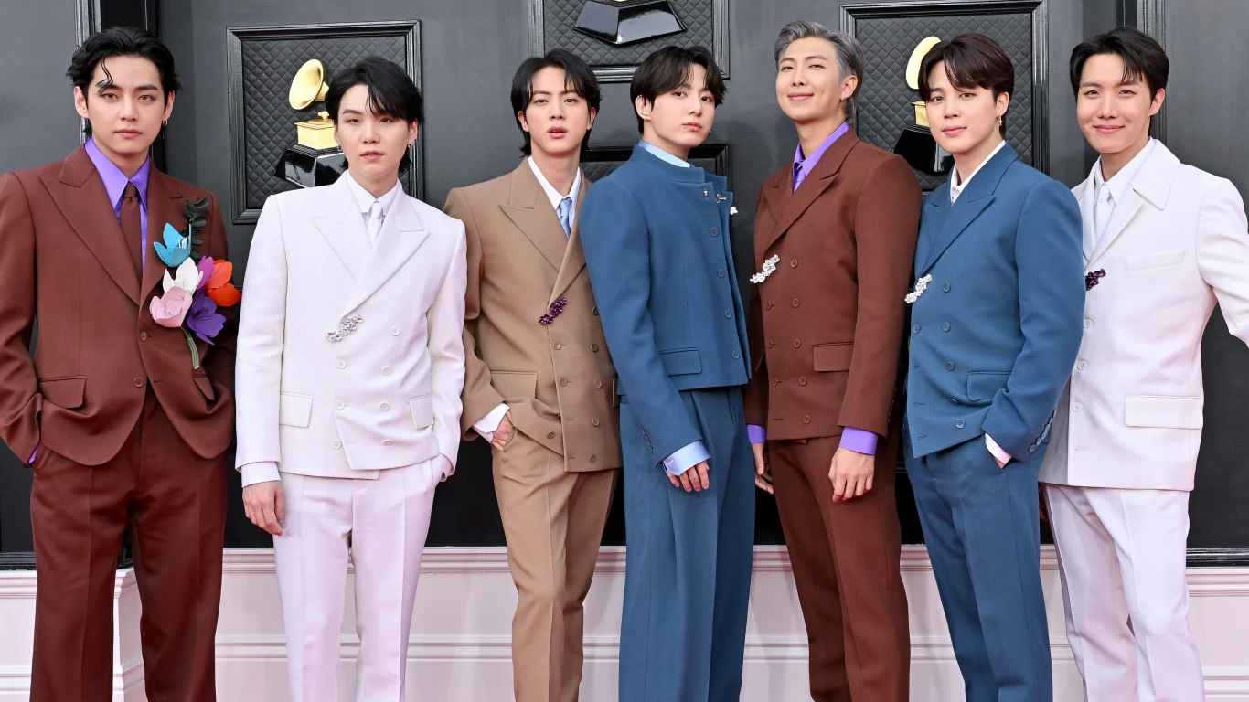 BTS (od lewej: V, Suga, Jin, Jungkook, RM, Jimin i J-Hope) Siedmiu elegancko ubranych mężczyzn w kolorowych garniturach pozuje grupowo na tle ściany z motywami muzycznymi, prezentując uśmiechy i pozy przed obiektywem.