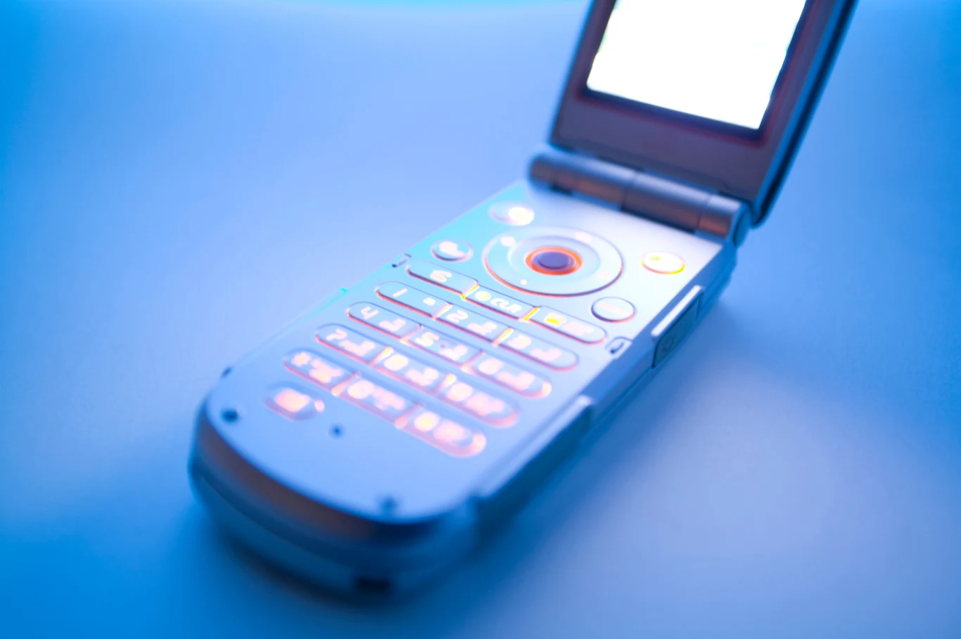 Cyfrowy detoks wśród młodzieży. Jak pokolenie Z walczy z uzależnieniem? Telefon komórkowy z klapką w srebrnej obudowie, podświetlana klawiatura na niebieskim tle, ekran rozjaśniony światłem.