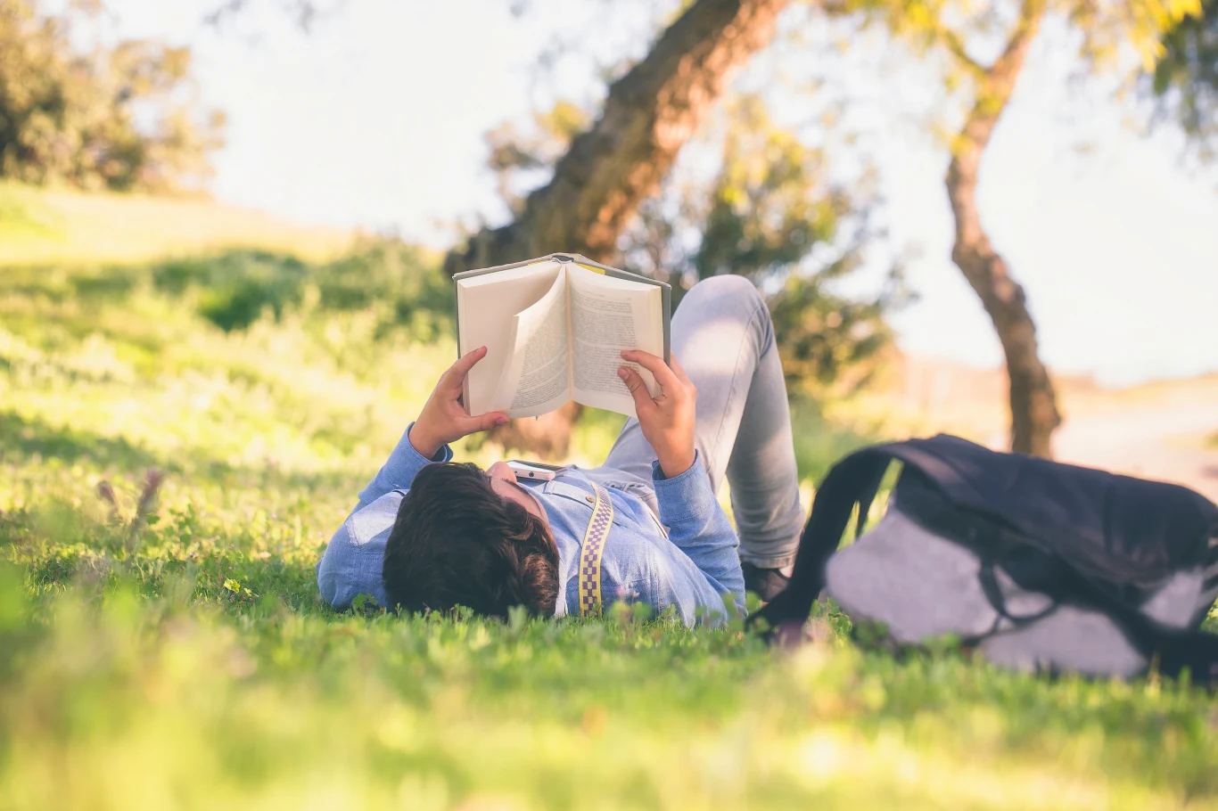 Wielu nastolatków wybiera czytanie książki zamiast scrollowania TikToka Osoba leżąca na trawie w parku czyta książkę, obok leży czarny plecak, w tle drzewa i promienie słońca.