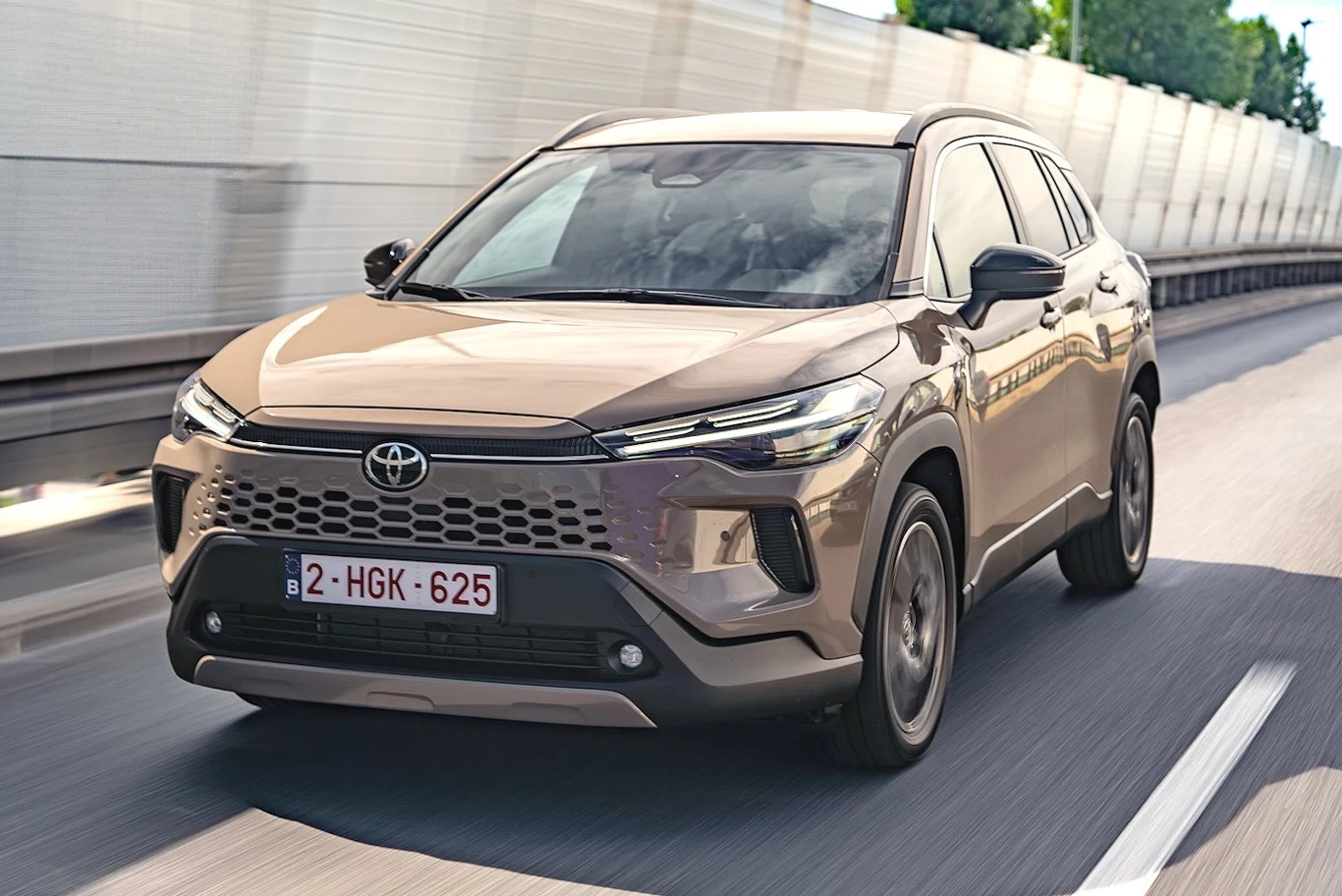Brązowy samochód Toyota jadący po asfaltowej drodze w otoczeniu betonowych ekranów dźwiękochłonnych, widok z przodu pojazdu, widoczna belgijska tablica rejestracyjna oraz charakterystyczny grill.