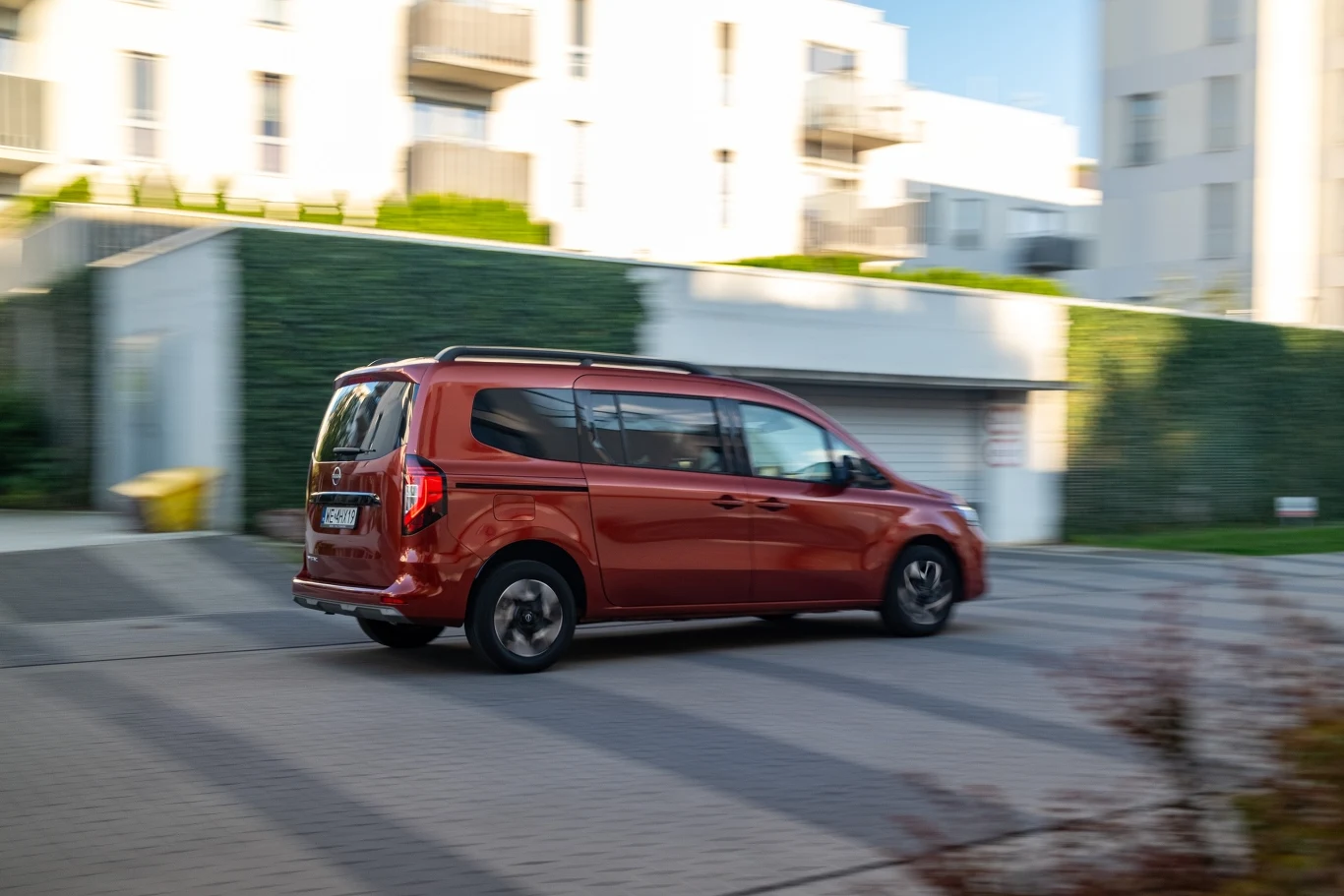 Czerwony samochód typu van poruszający się po osiedlu z nowoczesną zabudową, w tle białe budynki i zadbana zieleń ogrodzenia.