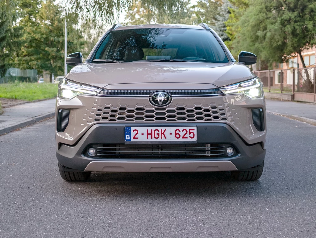 Toyota Corolla Cross otrzymała zupełnie nowy przód. Zniknął m.in. wyraźnie nakreślony grill, ktory teraz jest integralną częścią zderzaka Nowoczesny SUV marki Toyota o jasnobeżowym kolorze, widoczny z przodu na tle miejskiej ulicy, z wyraźnie widoczną belgijską tablicą rejestracyjną, światła przednie włączone, otoczenie zielone i miejskie.