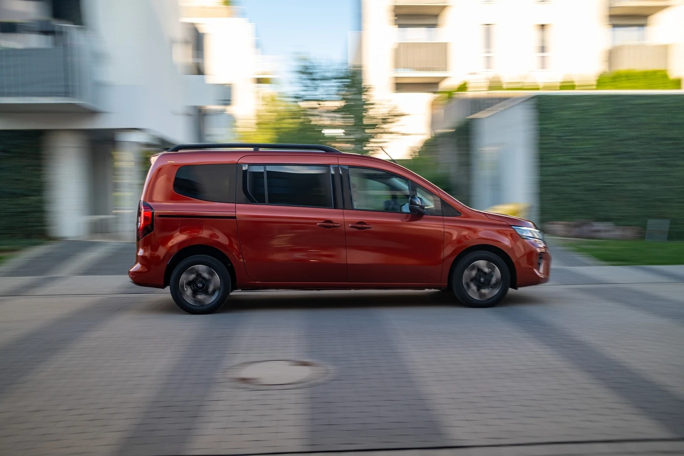 Czerwony minivan jadący ulicą w nowoczesnym osiedlu mieszkaniowym, pojazd w ruchu na tle rozmytych budynków i zieleni.