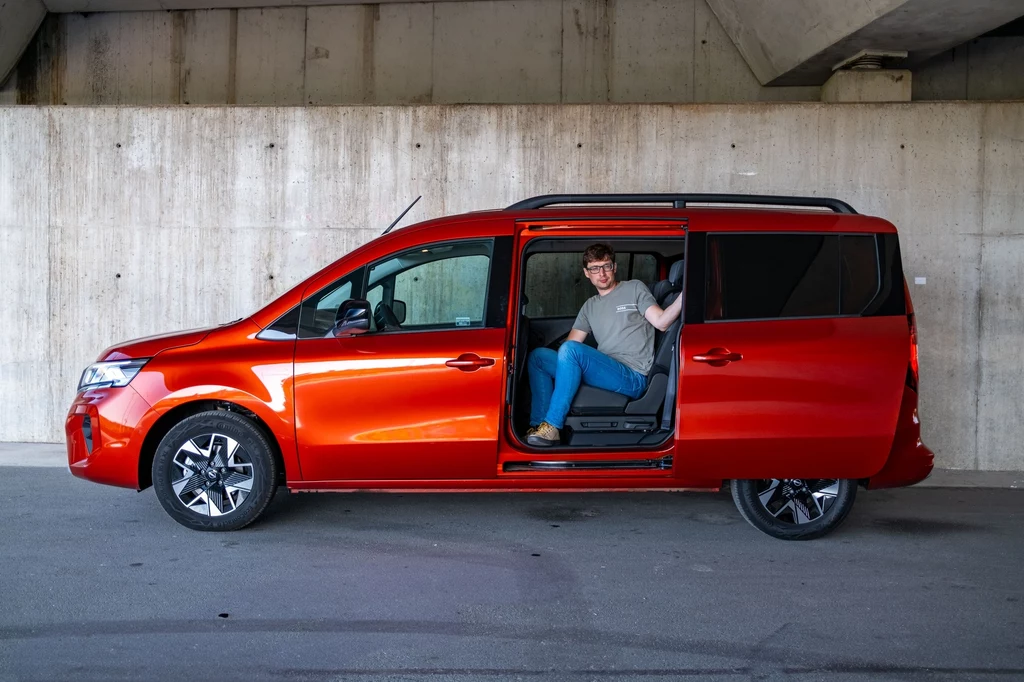 Kto używał, ten wie - na ciasnych parkingach nie ma nic lepszego niż przesuwane drzwi boczne Czerwony minivan zaparkowany pod betonowym wiaduktem, z rozsuwanymi drzwiami bocznymi, w których siedzi uśmiechnięty mężczyzna w okularach, machający ręką.