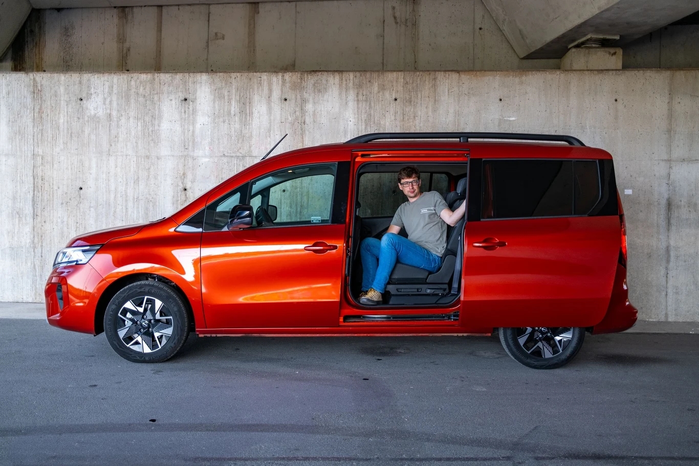 Czerwony minivan zaparkowany pod betonowym wiaduktem, z rozsuwanymi drzwiami bocznymi, w których siedzi uśmiechnięty mężczyzna w okularach, machający ręką.