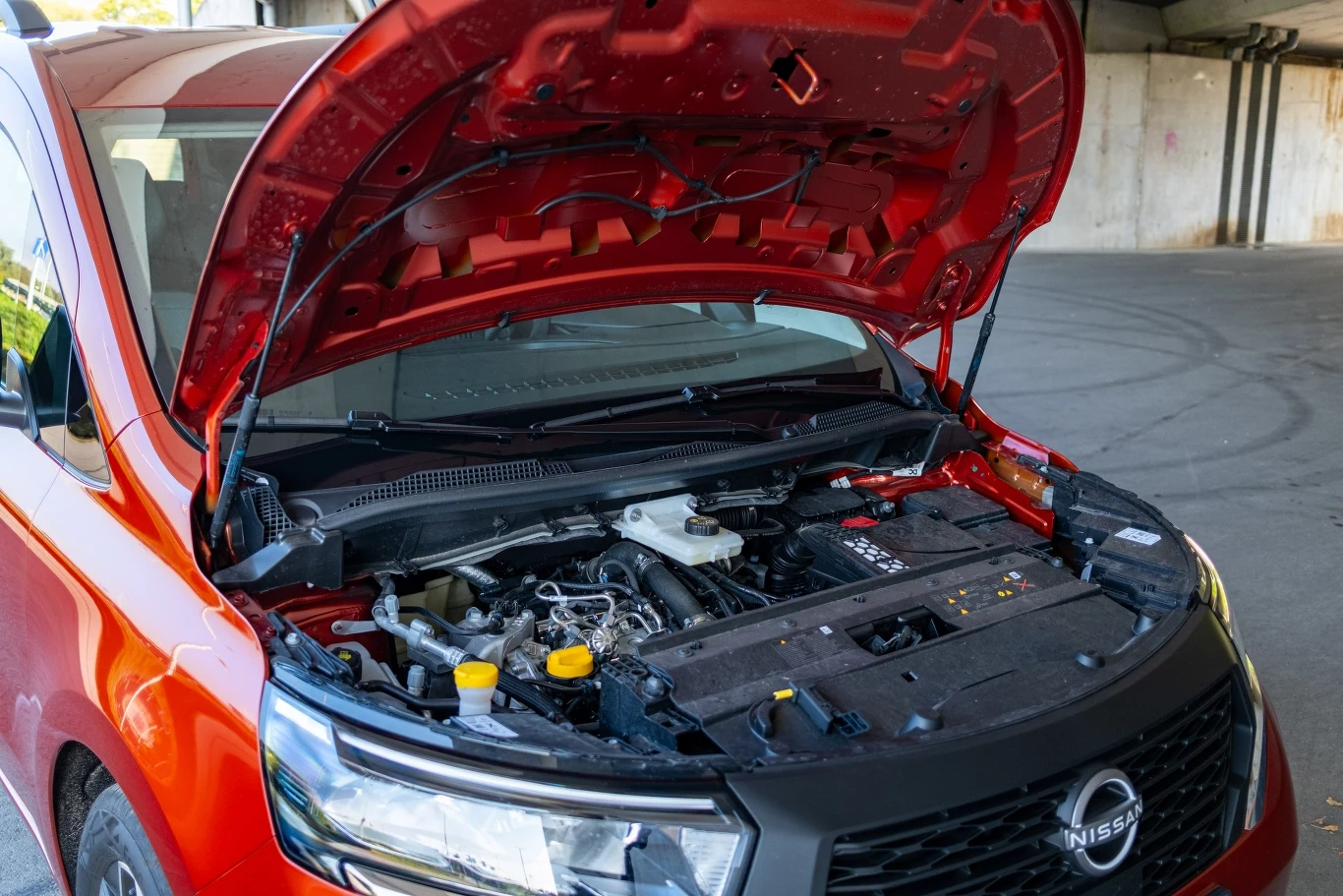 Czerwony samochód marki Nissan z otwartą maską, ukazujący szczegóły silnika oraz podzespołów pod maską na tle betonowej konstrukcji parkingu.