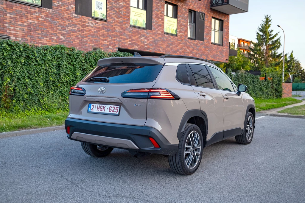 Wymiary Toyoty Corolli Cross po kuracji odmładzającej nie uległy zmianie Srebrny SUV marki Toyota Corolla Cross stoi zaparkowany na ulicy, w tle widoczny fragment nowoczesnego budynku z cegły oraz zielona roślinność przy płocie.