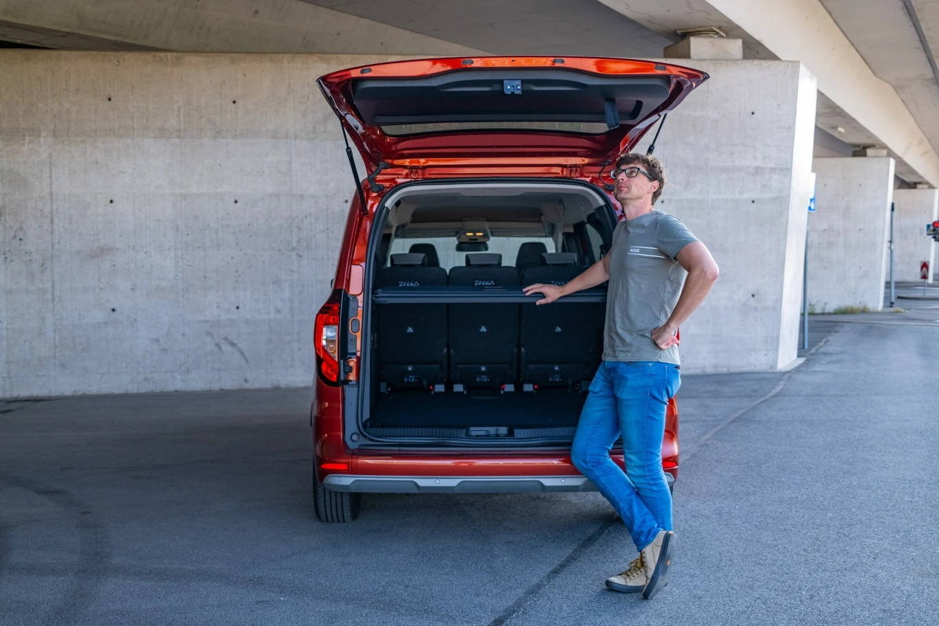 Osoba w okularach stoi oparta o czerwony samochód z otwartym bagażnikiem, zaparkowany pod betonowym wiaduktem, wnętrze auta pokazuje trzeci rząd siedzeń.